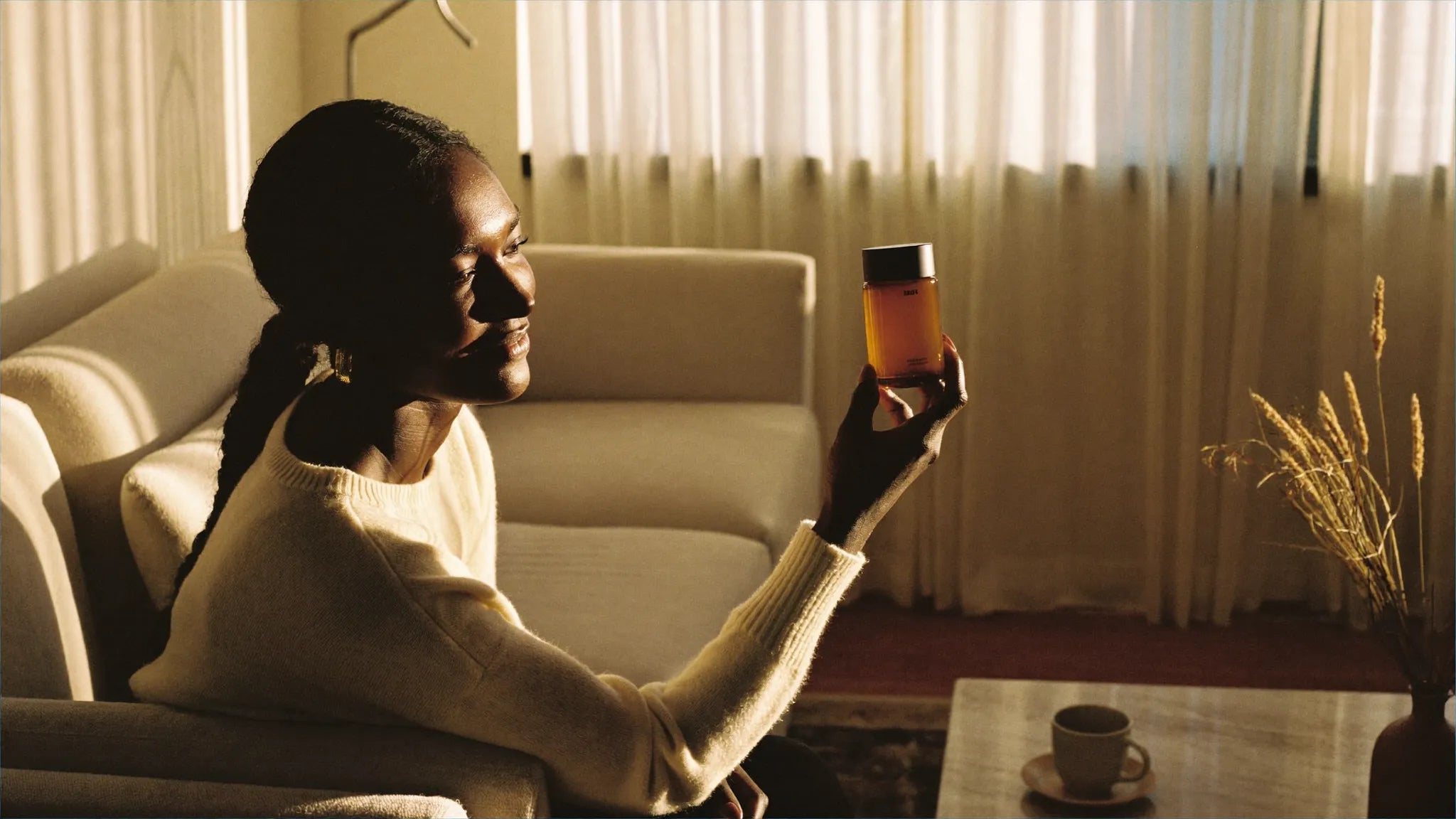 A woman in a sun-drenched living room reading the label of a honey-amber glass bottle, golden hour light